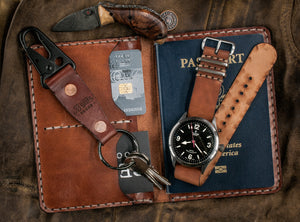 brown leather passport wallet with two card pockets and internal sleeve next to keychain, watch strap, and knife