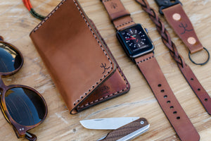 overlay of whiskey cordovan leather with brown interior four pocket vertical wallet next to watch, leather bracelet, and keychain