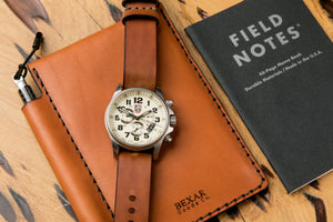 detail of back exterior of tan colored leather notebook wallet with pen sleeve next to analog watch