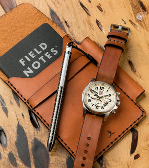 tan colored leather notebook wallet with pen sleeve next to analog watch
