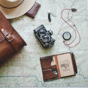 leather notebook wallet with two card pockets and pen sleeve next to compass, satchel, and camera
