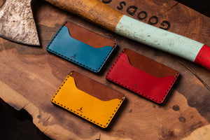 three wallets with two pocket with one center divider shown in yellow, red, blue leather with brown interior next to axe