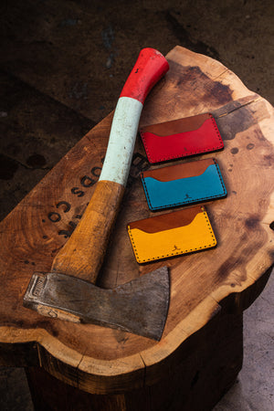 three wallets with two pocket with one center divider shown in yellow, red, blue leather with brown interior next to axe