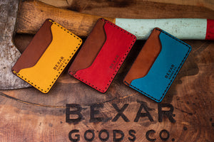 three wallets with two pocket with one center divider shown in yellow, red, blue leather with brown interior next to axe