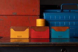 three wallets with two pocket with one center divider shown in yellow, red, blue leather with brown interior next to toolbox