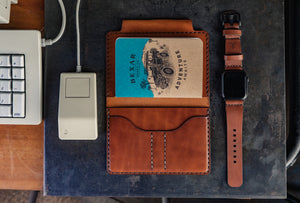overview of desk items next to computer showcasing brown leather  notebook wallet with card sleeves and passport sleeve next to smart watch