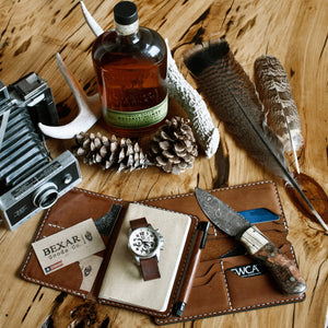brown leather long wallet with eight card pockets and two cash sleeves next to passport wallet, camera, feathers, and watch