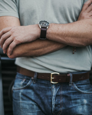 Adult male crosses arms showcasing a stainless watch with dark brown leather strap and dark brown belt.