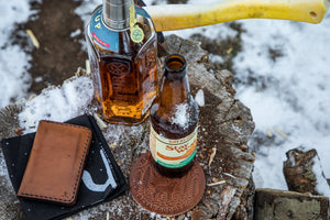 folded brown leather four pocket vertical wallet next to leather coaster, drinks, and axe in snow