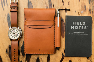 tan colored leather notebook wallet with pen sleeve next to analog watch