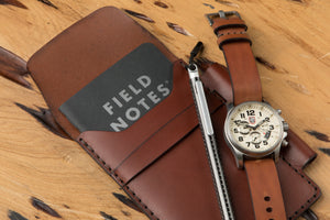 medium brown leather notebook wallet with pen sleeve next to analog watch