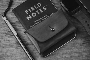 green leather folding card wallet with brass stud closure next to notebook and pen