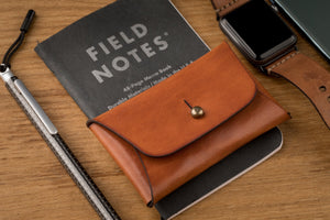 brown leather folding card wallet with brass nipple closure next to watch, keychain, and pen