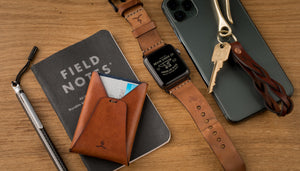 brown leather cardholder next to array of every day carry items- watch, leather keychain, and phone