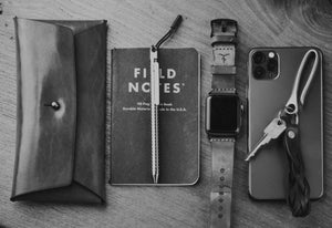 green leather folding card long wallet with brass stud closure adjacent to notebook, watch strap, keychain