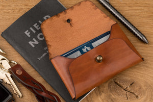 open view of brown leather folding card wallet with brass nipple closure next to watch, keychain, and pen