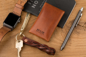 rear view of brown leather folding card wallet with brass nipple closure next to watch, phone, and pen