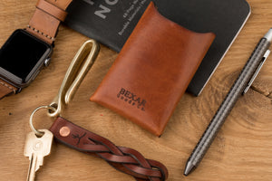 rear view of brown leather cardholder next to array of every day carry items- watch, leather keychain, and phone