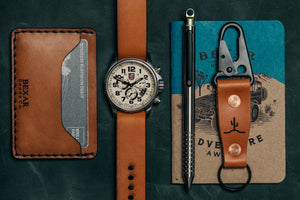 overview of brown leather two pocket wallet surrounded by analog watch, pen, and keychain